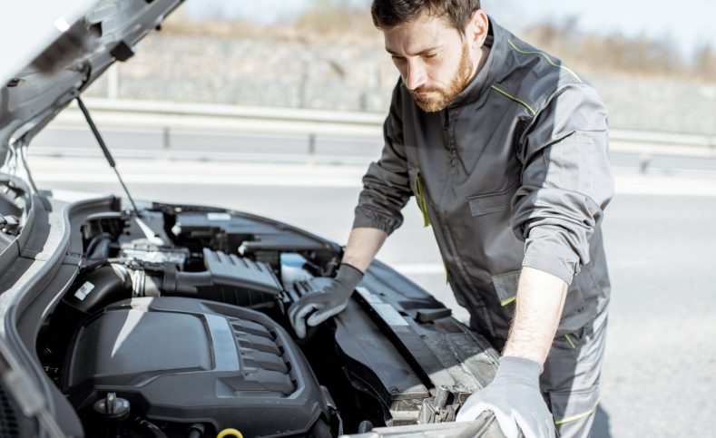 Handsome auto mechanic or road assistance worker in uniform repairing engine of the broken car on the road