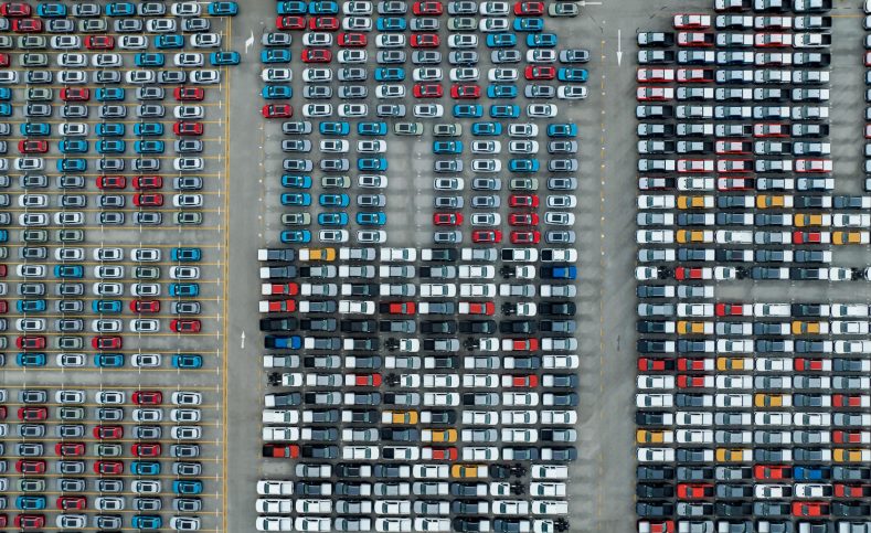 Aerial view of new cars stock at factory parking lot. Above view cars parked in a row. Automotive industry. Logistics business. Import or export new cars at warehouse. Big parking lot at port terminal